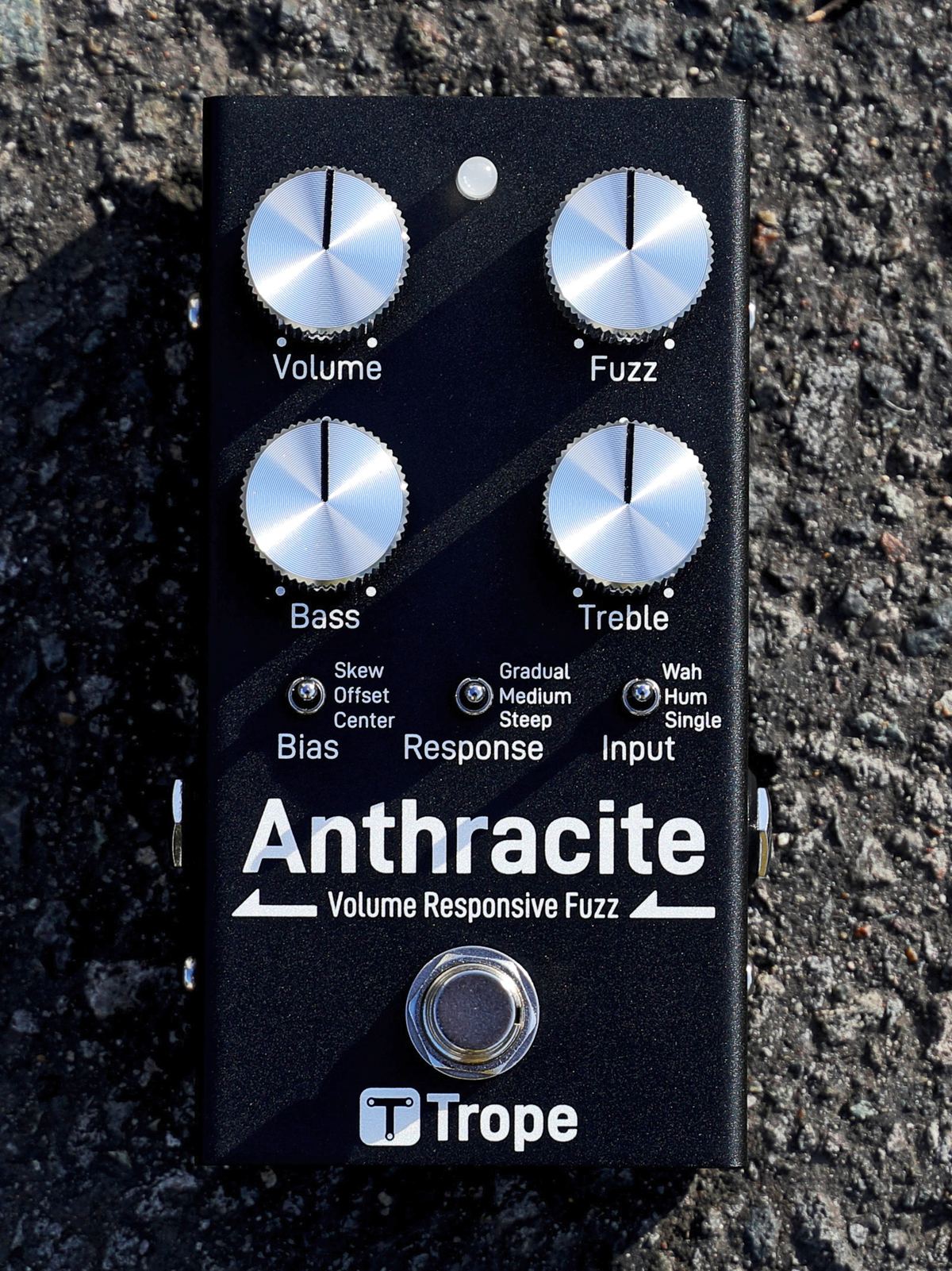 アスファルトの上に置いた Anthracite v1.0 を真上から見たところ。黒い筐体にアルミのつまみ 4 個（Volume / Fuzz / Bass / Treble）、トグルスイッチ 3 個（Bias / Response / Input）、フットスイッチが並ぶ