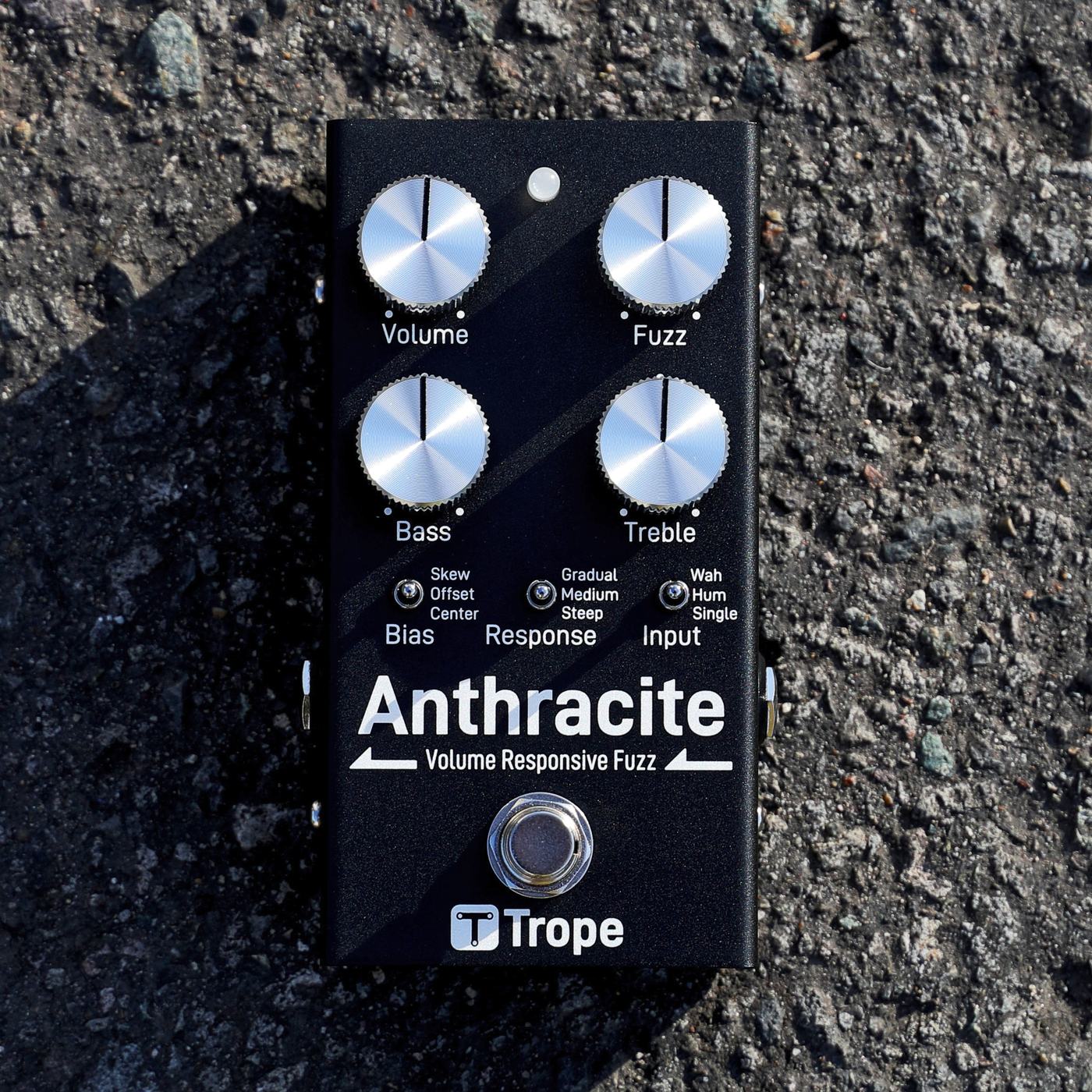 アスファルトの上に置いた Anthracite v1.0 を真上から見たところ。黒い筐体にアルミのつまみ 4 個、トグルスイッチ 3 個、フットスイッチが並ぶ